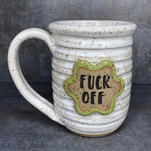 May include: White ceramic mug with a green and brown accent featuring the phrase "F*ck Off" in black lettering.