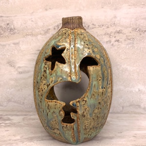 May include: A ceramic vase with a face design. The vase is oval-shaped with a textured surface and a star-shaped cutout. The color is a mix of brown, green, and blue, with a textured finish. The top has a ribbed texture.