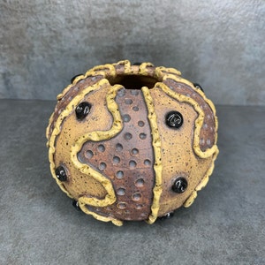 May include: A round ceramic vase with a textured brown and yellow design. The vase has a wide opening and is decorated with black dots.