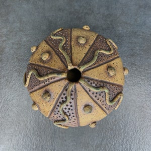 Pode incluir: Um vaso de cerâmica com um esmalte marrom e amarelo salpicado. O vaso tem um design único com pontos em relevo e linhas verdes que se assemelham a um ouriço-do-mar.