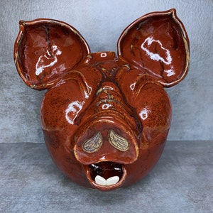 May include: A ceramic piggy bank with a red glaze and a brown snout. The piggy bank has a wide, open mouth with white teeth.