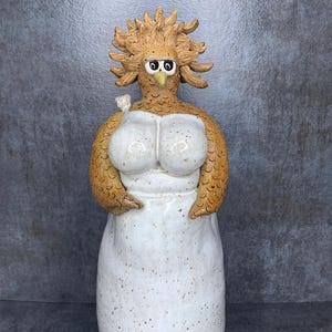 以下が含まれることがあります： 鶏の頭をつけた女性のセラミック彫刻で、白いドレスに茶色のアクセントが施されています。彫刻は風変わりでユーモラスなスタイルです。