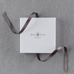 Puede incluir: Una caja blanca con una cinta gris atada alrededor. La caja tiene el texto "Jenny Grace Jewellery" impreso en ella.