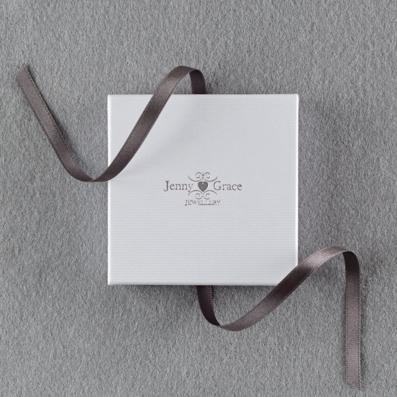 Puede incluir: Una caja blanca con una cinta gris atada alrededor. La caja tiene el texto "Jenny Grace Jewellery" impreso en ella.