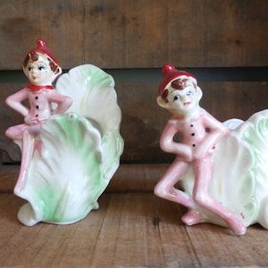 May include: Two vintage ceramic elf figurines, each holding a green cabbage leaf planter. The elves are wearing pink outfits and red hats.