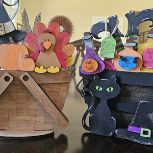 May include: Two wooden decorative baskets, one with a Thanksgiving theme and the other with a Halloween theme. The Thanksgiving basket features a turkey, pumpkins, and a pie. The Halloween basket features a black cat, a witch's hat, and potion bottles with labels that say "Pick Your Poison", "Bat Wings", "Rat Tails", and "Eye of Newt".
