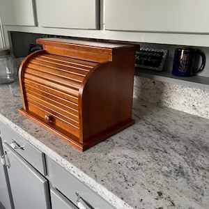 May include: A medium-toned wooden bread box featuring a roll-top tambour door with a small round knob. The rectangular storage container has a flat top surface and a solid base, designed for countertop use to store baked goods in a kitchen.