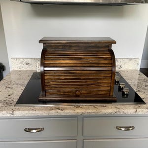 May include: A dark brown wooden bread box with a roll-top door featuring horizontal slats and a small round knob. The rectangular storage container has a flat top surface and a solid base, showcasing a visible wood grain finish throughout the structure.