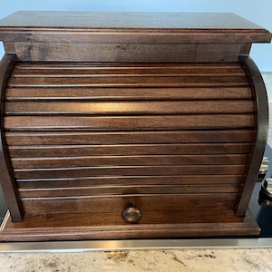May include: A dark brown wooden bread box featuring a roll-top door with horizontal slats and a small round knob. The rectangular container has a flat top surface and a solid base, providing a storage solution for kitchen items.