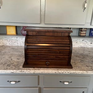 May include: A dark brown wooden bread box with a roll-top door and a small circular knob. The bread box features horizontal slats across the curved front panel and a flat top surface, positioned on a speckled granite countertop.