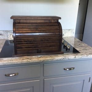 May include: A dark brown wooden bread box featuring a roll-top door with horizontal slats and a small round knob. The rectangular container has a flat top surface and a sturdy base, showcasing a polished wood grain finish throughout the structure.