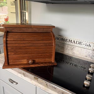 May include: A medium-brown wooden bread box featuring a roll-top door with a small round knob. The rectangular storage container has a flat top surface and a slatted front design, constructed from wood with a visible grain pattern throughout.