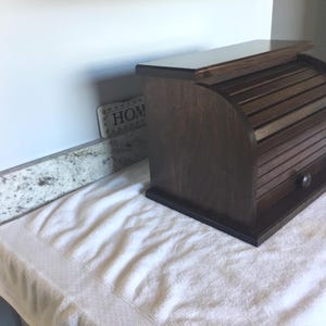 May include: A dark brown wooden bread box with a roll-top door and a small knob. The box is rectangular with rounded sides and sits on a white towel. A small sign with the word "HOME" is visible in the background.