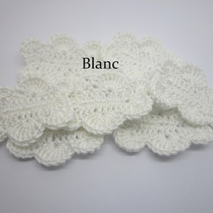 Op de afbeelding: Een close-up van een stapel witte gehaakte bloemenversieringen. Het woord "Blanc" staat in zwarte tekst bovenop de stapel.