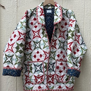 May include: A quilted jacket featuring a repeating star pattern in red, green, and white. The garment has a notched collar, long sleeves with dark blue floral cuffs, and two front patch pockets. A label inside reads Designs by Studio C.