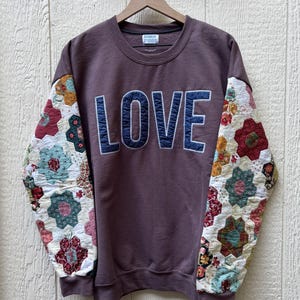 May include: A dark mauve crewneck sweatshirt featuring the word LOVE in large, dark blue quilted letters across the chest. The long sleeves are constructed from a white fabric with a multi-colored floral hexagon patchwork quilt pattern.