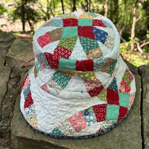 May include: A handmade patchwork sun hat with a wide brim. The hat features a variety of fabric squares in red, green, pink, and floral patterns, sewn together in a circular design. The brim is trimmed with a dark blue fabric.