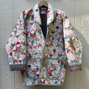 May include: A quilted jacket with a patchwork design in various colors, including red, teal, and orange. The jacket has a collar, pockets, and a brown polka dot trim. The label reads "DESIGNED BY STUDIO C."