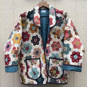 May include: A quilted jacket featuring a patchwork of hexagonal floral patterns in shades of red, yellow, blue, and cream. The garment has a notched collar, long sleeves with teal cuffs, two front patch pockets, and a teal interior lining with a label.
