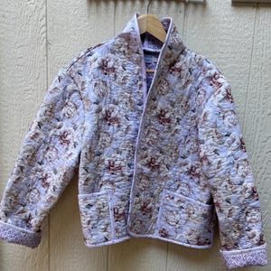 May include: A quilted jacket featuring a light purple base with a repeating floral pattern in shades of pink, cream, and brown. The jacket has a shawl collar, long sleeves with folded cuffs, and two front patch pockets with light purple trim.