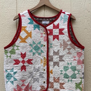 May include: A quilted vest featuring a patchwork star pattern in red, teal, yellow, green, and brown on a white background. The vest has a red polka-dot trim, five gold-tone metal buttons, and a label reading Designs by Studio C at the neckline.