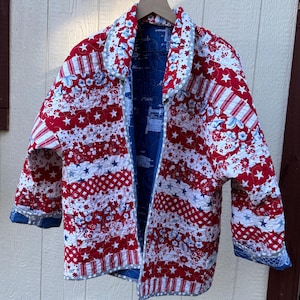 May include: A quilted jacket with a patriotic theme, featuring red, white, and blue patterns. The jacket has a reversible design with a blue interior and a mix of star and stripe patterns on the exterior. The collar and cuffs are trimmed with a contrasting fabric.
