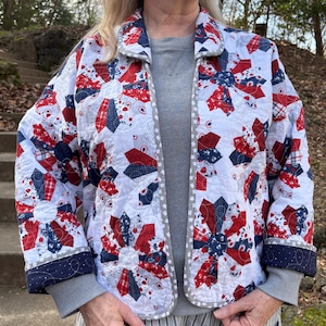 May include: A quilted jacket in a patriotic color scheme of red, white, and blue. The jacket features a floral pattern and a gray trim. The cuffs and collar are navy blue. The jacket is open in the front.