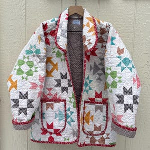 May include: A quilted jacket with a white background and colorful star patterns in red, orange, green, and blue. The jacket has a red trim, two pockets, and a brown interior. The label reads "Designed by Studio C".
