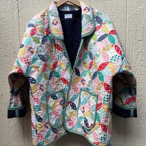 May include: A quilted jacket featuring a colorful patchwork pattern of overlapping petal shapes in red, yellow, green, and blue floral prints. The jacket has a notched collar, two front pockets, and a dark blue interior lining with white dots.