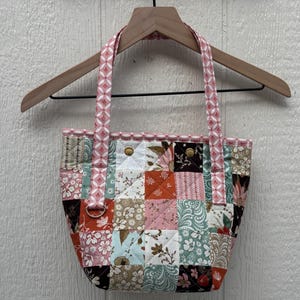 May include: A patchwork tote bag with a variety of floral patterns in shades of pink, orange, blue, and brown. The bag has a quilted design and pink handles with a geometric pattern. The bag is hanging on a wooden hanger.