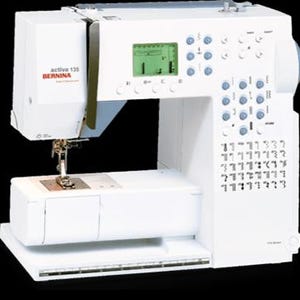以下が含まれることがあります： デジタルディスプレイと多数のボタンを備えた白いBernina Activa 135ミシン。ミシンには針、押え金、作業面があります。ブランド名とモデルが確認できます。