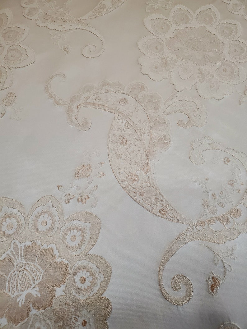 May include: Close-up of a white fabric with a beige floral pattern. The fabric has a raised texture and a delicate design.