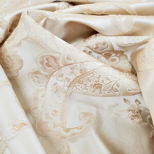 May include: Close-up of a cream-colored fabric with a raised, embossed paisley pattern in a slightly darker shade of cream.