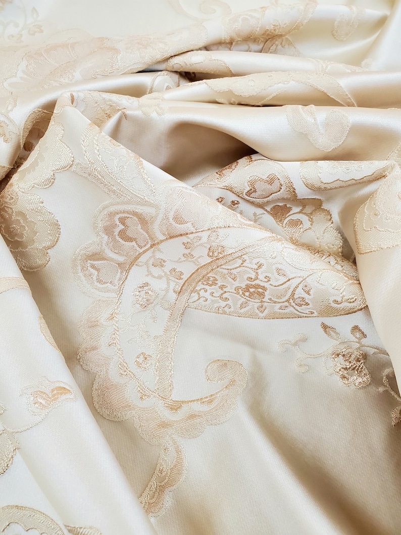 May include: Close-up of a cream-colored fabric with a raised, embossed paisley pattern in a slightly darker shade of cream.