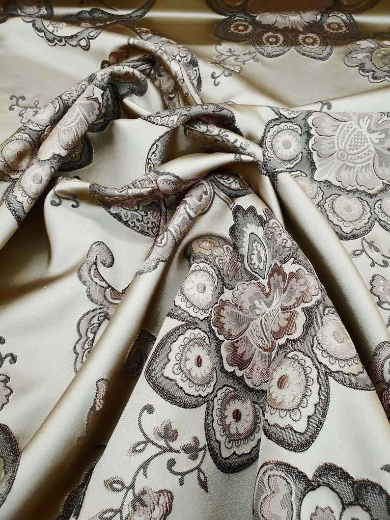 May include: A beige fabric with a brown and gray floral pattern. The fabric is folded and draped, showing the intricate details of the design.