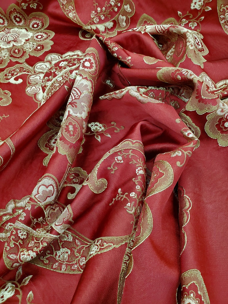 May include: Red fabric with a gold paisley pattern. The fabric is folded and draped, showing the texture and detail of the design.