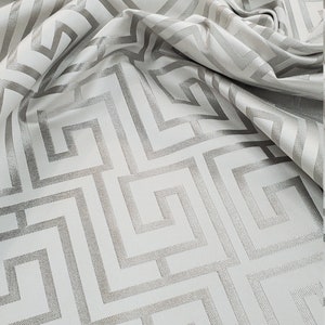 May include: White fabric with a silver geometric pattern. The pattern is a repeating design of squares and rectangles.