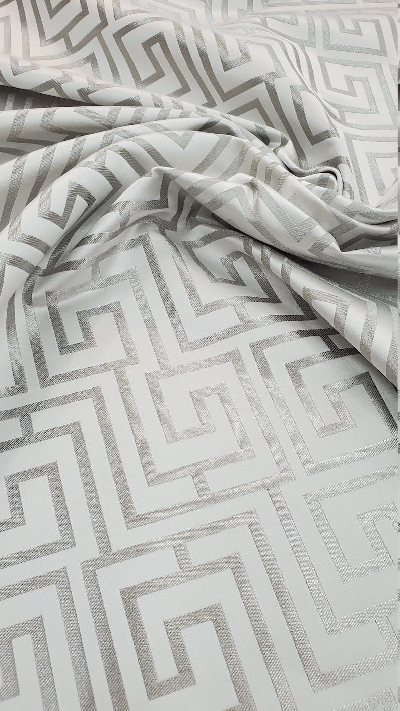 May include: White fabric with a silver geometric pattern. The pattern is a repeating design of squares and rectangles.