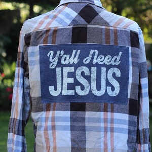 May include: A blue and white plaid flannel shirt with a blue patch on the back that says "Y'all Need Jesus" in white lettering.