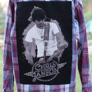 May include: A red and blue plaid flannel shirt with a black patch on the back. The patch features a photo of a man playing a guitar with the text "Chris Janson" in a circle with wings.