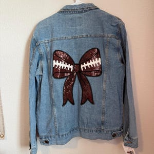 May include: Light blue denim jacket with a large sequined bow design on the back. The bow is a deep burgundy color with white stitching, resembling a football. The jacket has a classic button-up design with pockets and a collar.