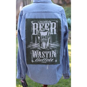 May include: A light blue denim shirt with a green patch on the back. The patch features a white graphic of a beer can with the text "Drinkin' Beer" and "Wastin' Bullets" with a rhinestone design. The beer can graphic also includes the text "Luke Bryan".