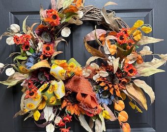 May include: A fall-themed wreath with sunflowers, orange and red flowers, and brown and green leaves. The wreath is decorated with a yellow and green ribbon with a sunflower pattern.