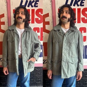 May include: Two people wearing olive green hooded jackets with multiple pockets and button closures. The jackets have a relaxed fit and are made of a durable fabric.