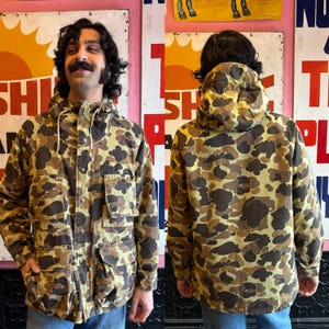 May include: A brown and tan camouflage jacket with a hood. The jacket has a zipper closure and multiple pockets.