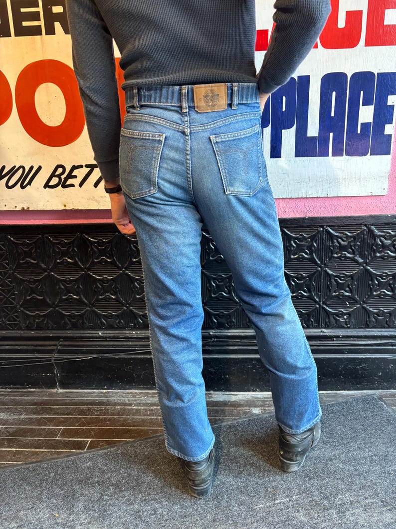May include: Light blue denim jeans with a classic five-pocket design and a brown leather belt. The jeans have a straight leg cut and are worn with black shoes. The background includes vintage signage.