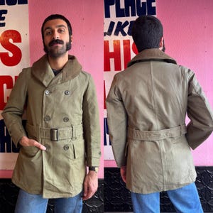 Puede incluir: Un abrigo verde oliva de doble botonadura con cuello marrón y cinturón a juego. El abrigo tiene dos filas de botones y dos bolsillos delanteros. El abrigo se muestra por delante y por detrás.