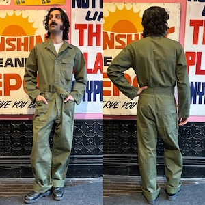 Può includere: Una tuta da lavoro verde con cerniera frontale, due tasche sul petto e due tasche laterali. La tuta ha una cintura e maniche lunghe.