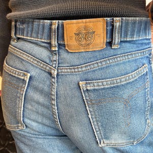 May include: Light blue denim jeans with a brown leather patch featuring the text "Great Western Garment Company" and a logo. The jeans have classic five-pocket styling and a straight leg fit.