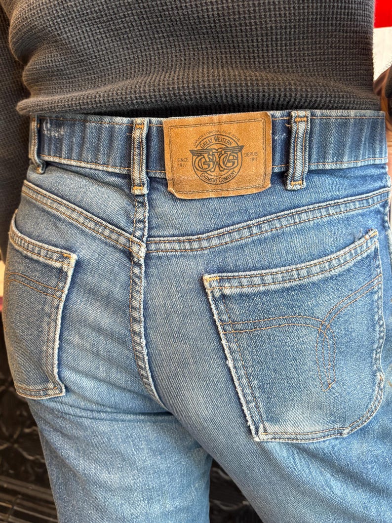 May include: Light blue denim jeans with a brown leather patch featuring the text "Great Western Garment Company" and a logo. The jeans have classic five-pocket styling and a straight leg fit.